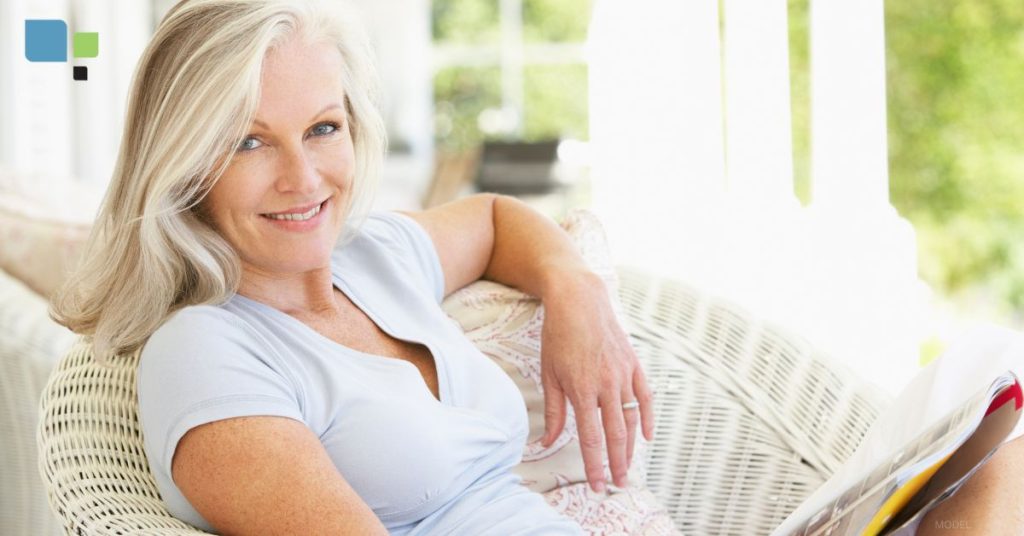 A woman with blonde hair is sitting outside smiling while sitting on a chair and wearing a white shirt. (model)